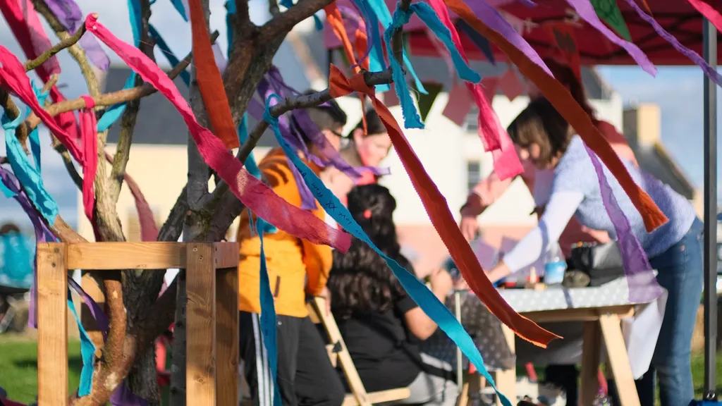 Ribbon image of Remembering Together D&G event, with memory ribbons fluttering in the wind and event visitors gating arround a table in the background.